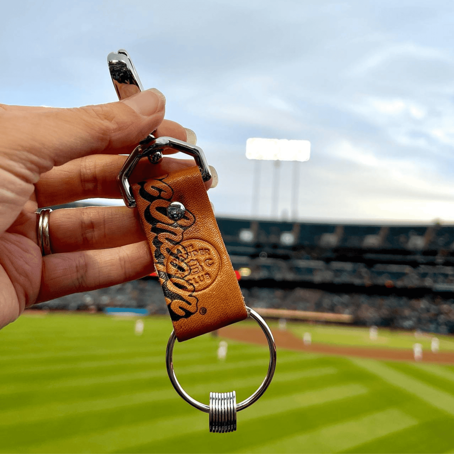 Close-up of a baseball glove Wanderchain keychain with orange leather strap, held against a baseball stadium background.