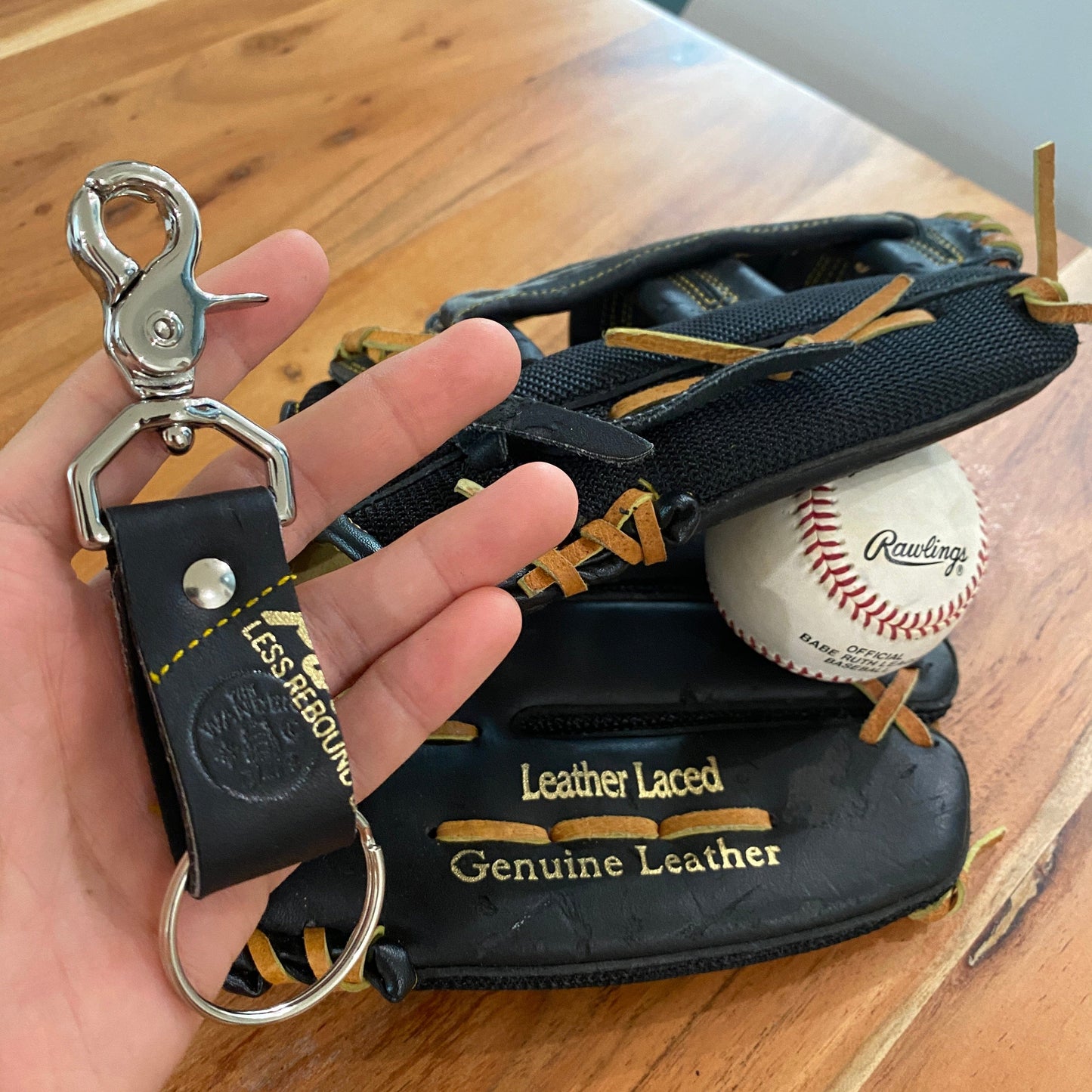 A close-up of a baseball glove with a Rawlings baseball, and a keychain with a metal clasp and leather strap, on a wooden surface.