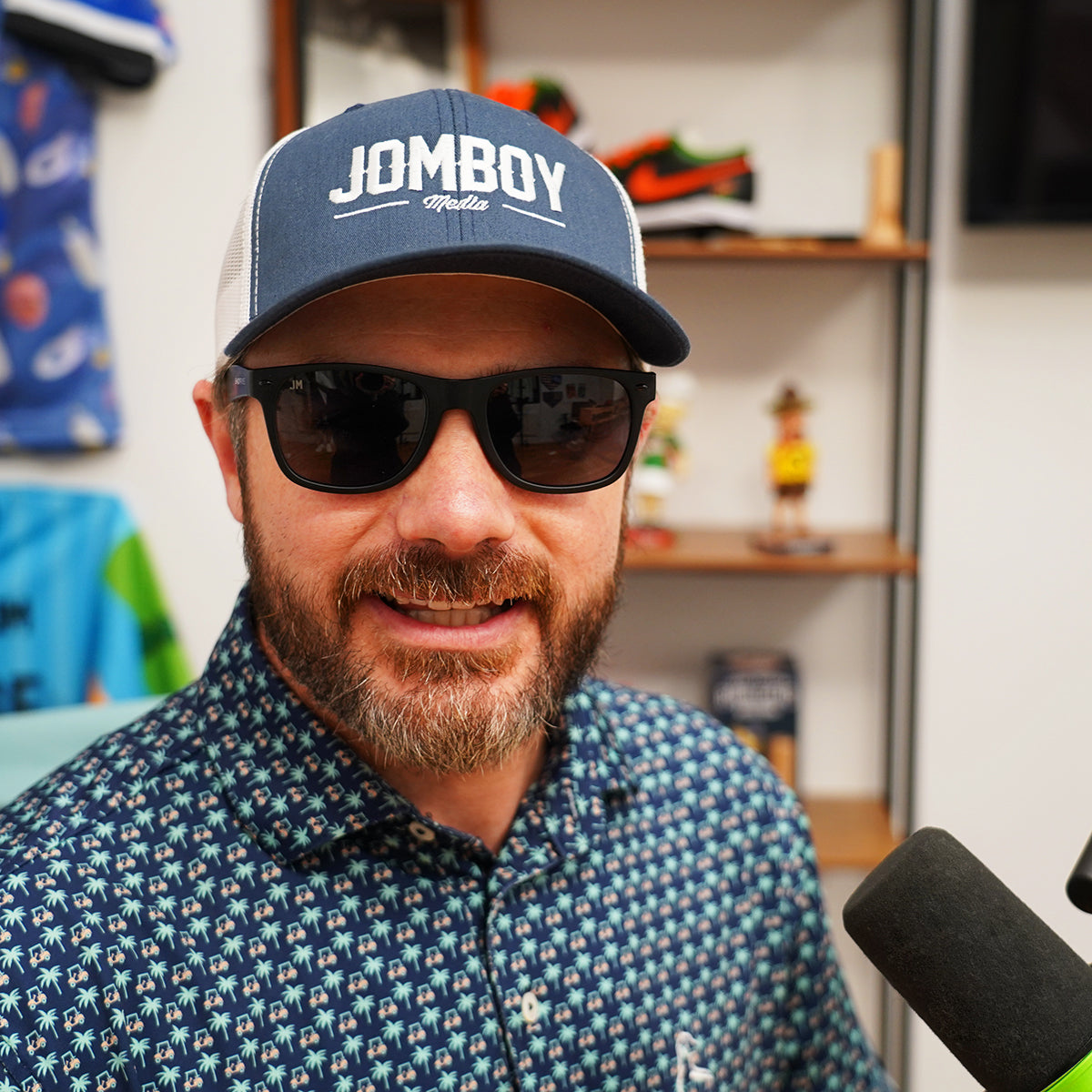 Man wearing black Shady Rays sunglasses, a blue "Jomboy Media" cap, and a patterned shirt, smiling indoors.