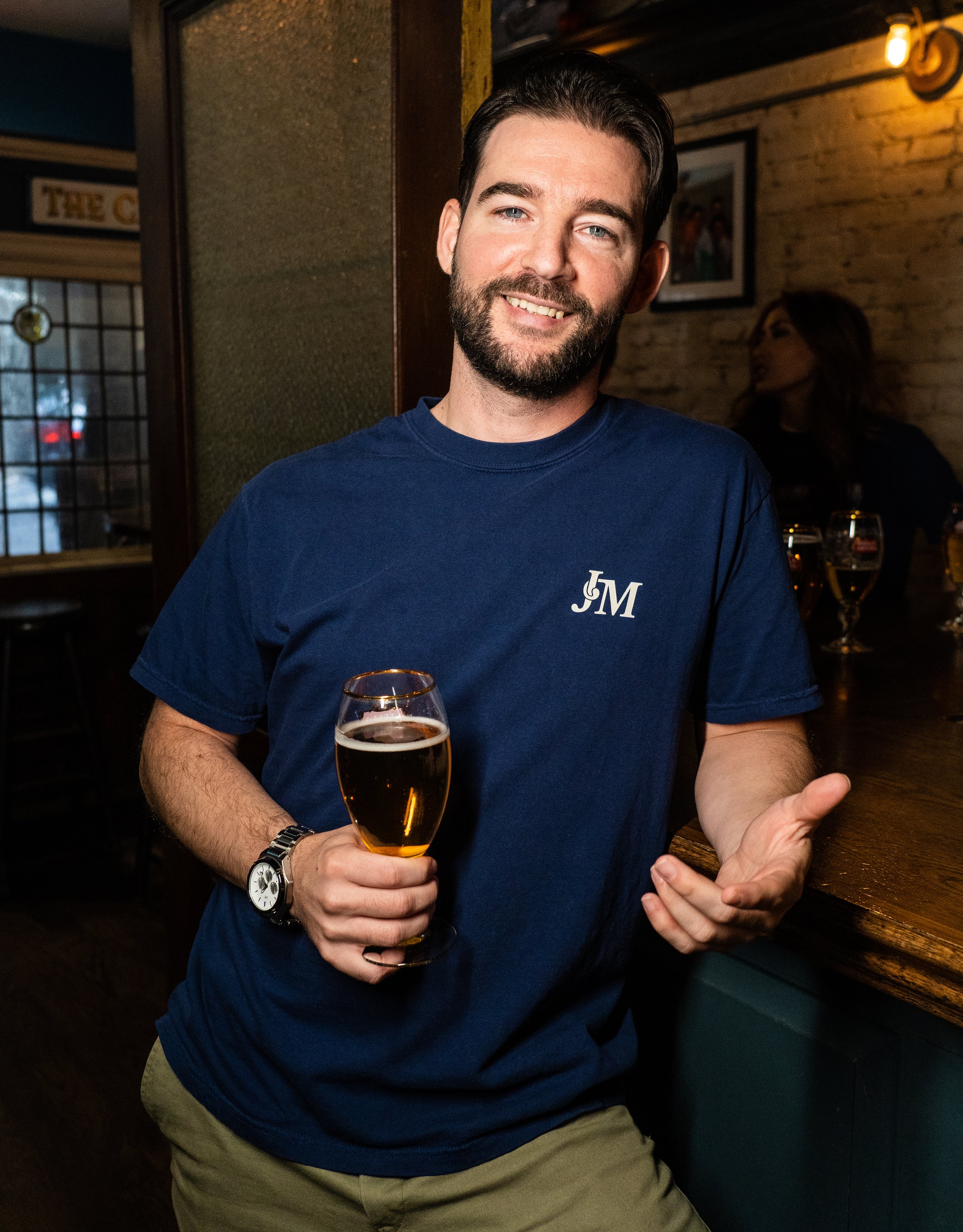 A smiling man with dark hair and a beard holding a glass of beer in a cozy bar, wearing a navy blue t-shirt with "J M" logo.