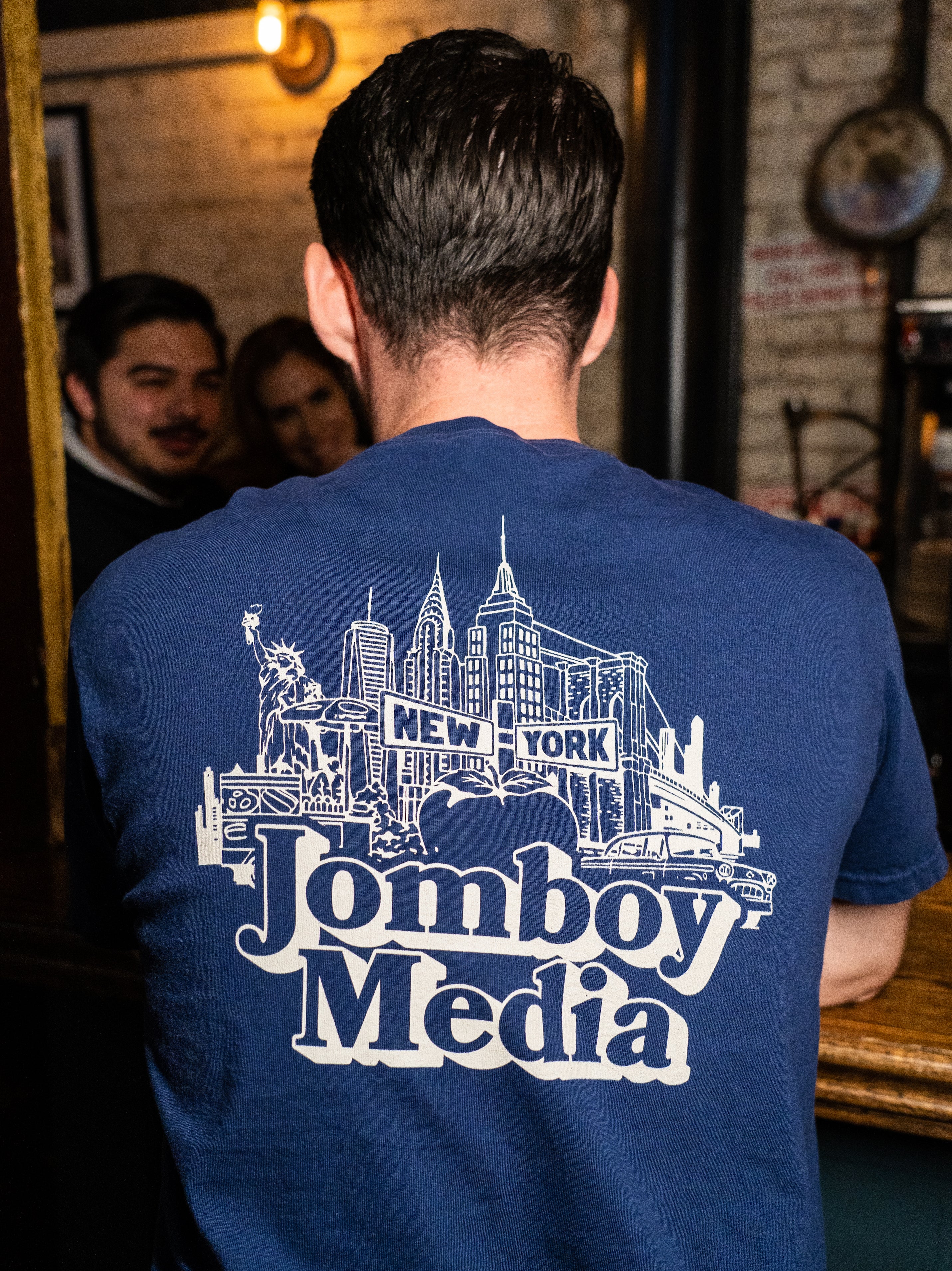 Back of a person wearing a blue T-shirt with a New York City skyline graphic and "Jomboy Media" text, in a cozy indoor setting.