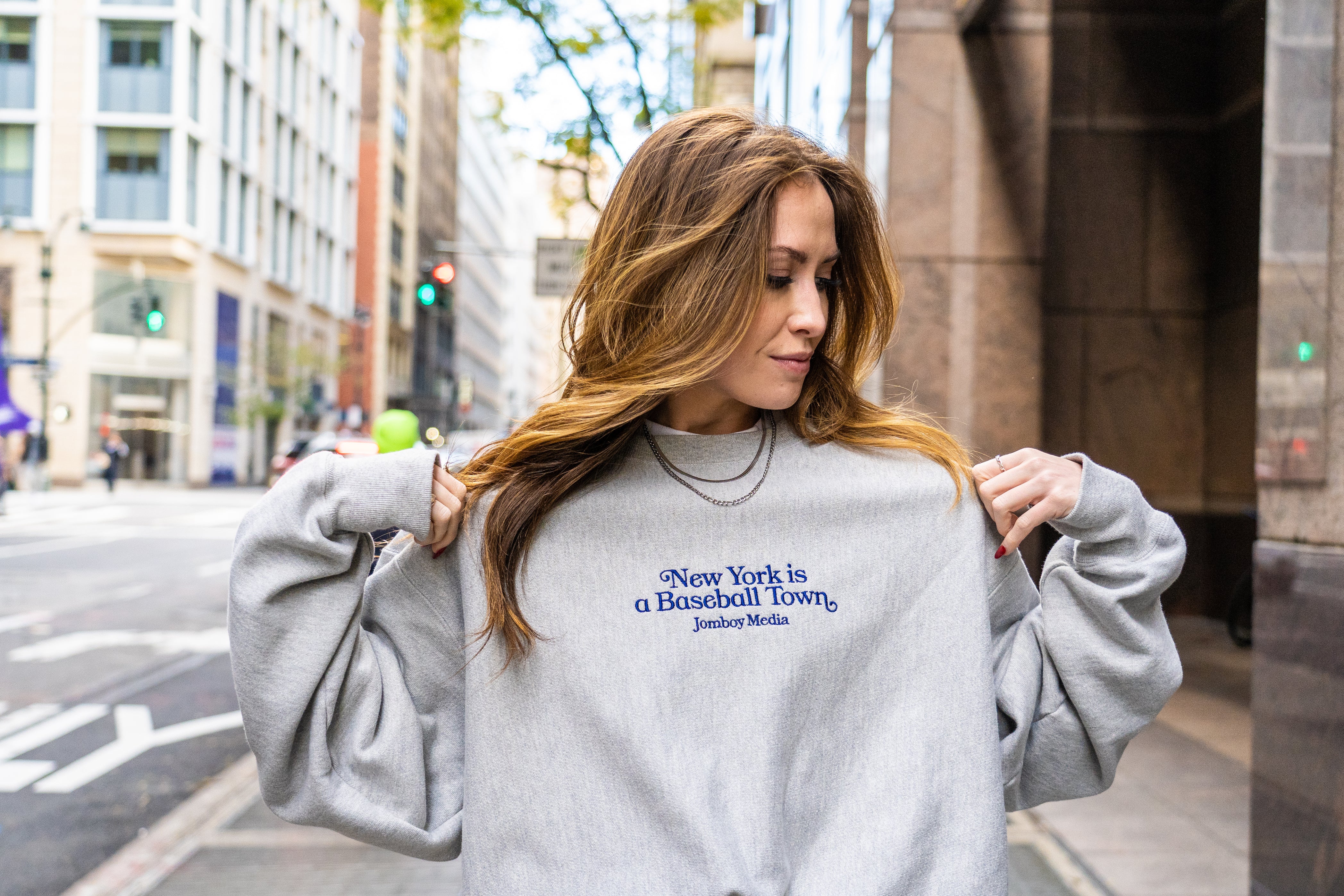 Woman wearing a light gray embroidered crewneck sweatshirt with "New York is a Baseball Town" text, standing on a city street.