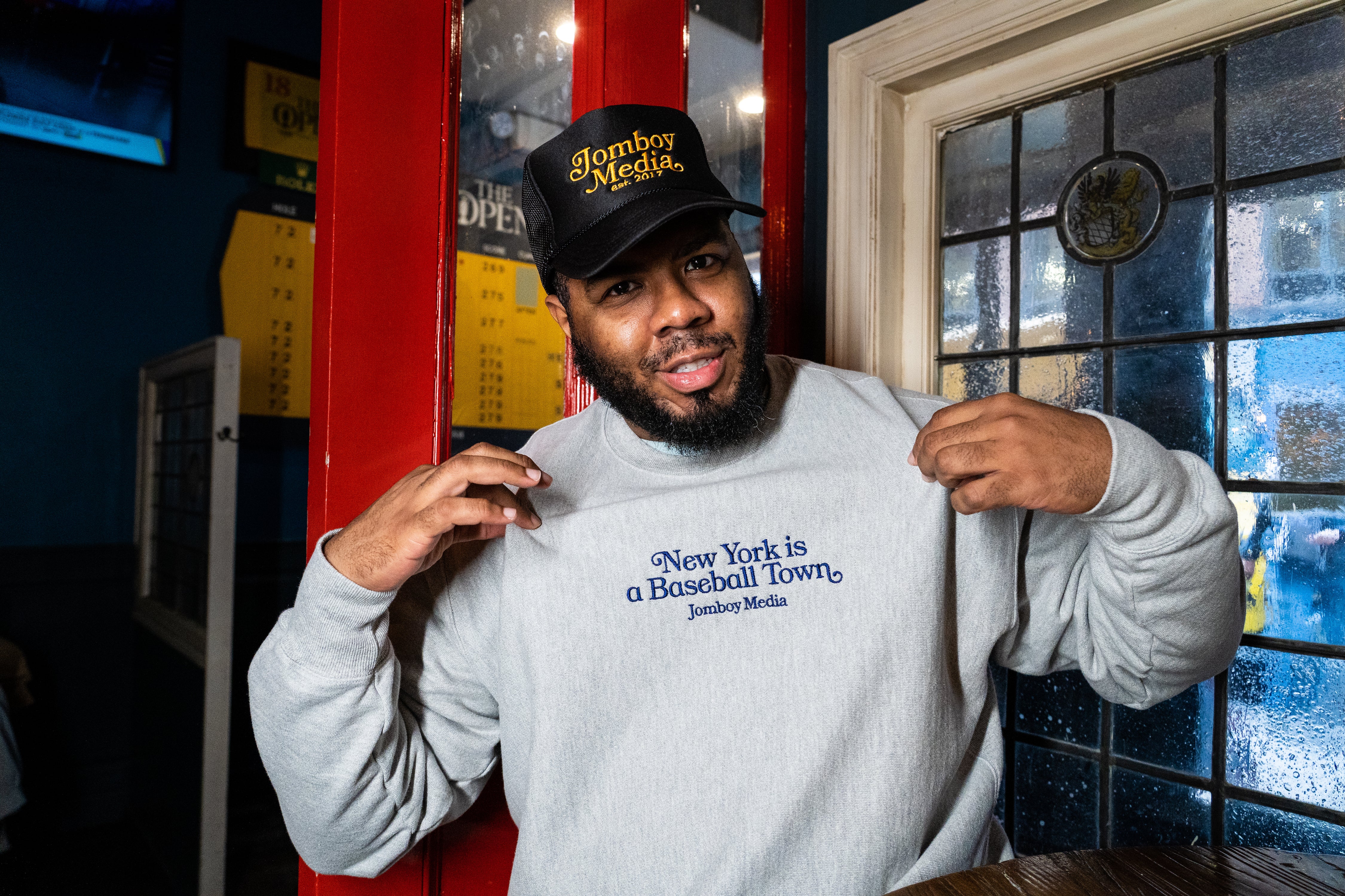 Man wearing a light gray sweatshirt with "New York is a Baseball Town" embroidered in blue, holding it out to show the design.