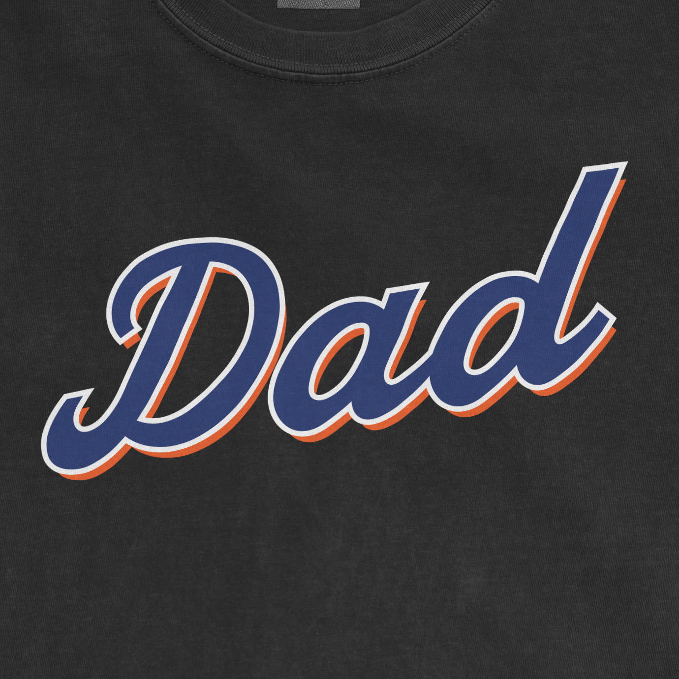 Close-up of a black T-shirt with "Dad" written in bold, blue script with orange and white outline, celebrating Queens baseball dads.