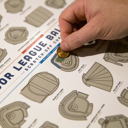 Close-up of a hand placing a coin on a Major League Baseball stadium scratch-off poster, featuring illustrated stadium layouts.
