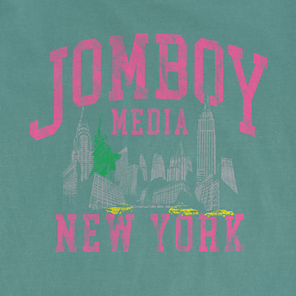 Comfort Colors tee featuring a vintage-inspired graphic of New York City landmarks, taxis, and "JOMBOY MEDIA 90's Edition" text in pink.