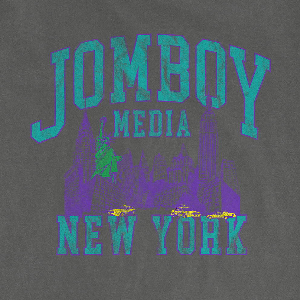 Gray Comfort Colors T-shirt featuring a neon-style graphic of New York City skyline, Statue of Liberty, taxis, and "JOMBOY MEDIA 90'S EDITION" text.