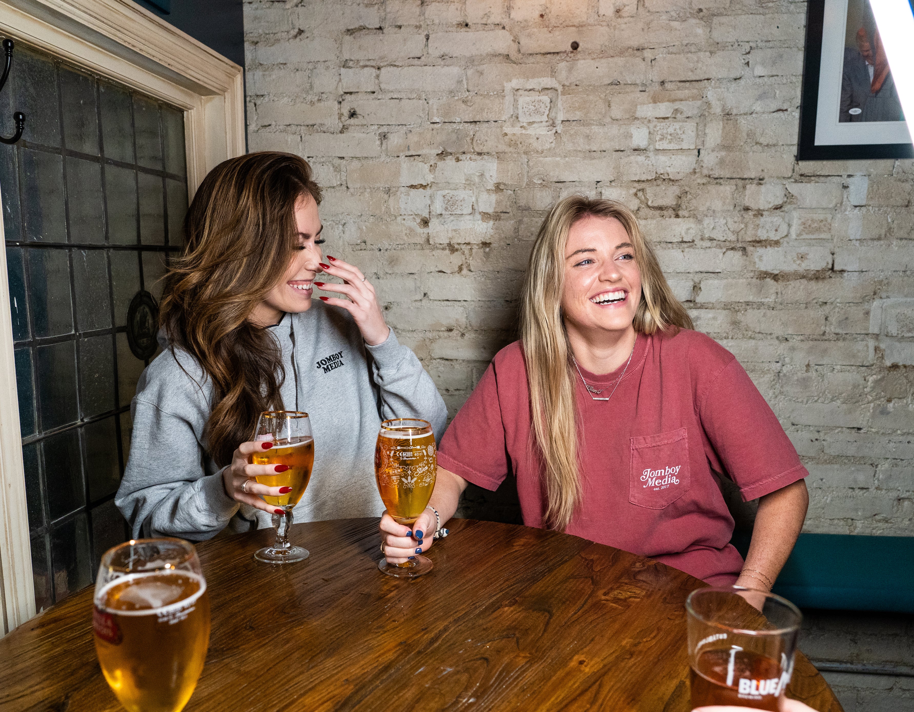 Two women laughing and enjoying drinks at a wooden table, wearing "Jomboy Media" branded apparel.